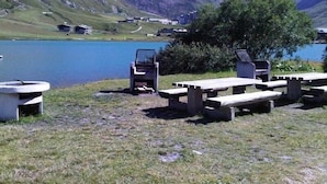 En picknickplats vid sjön med bänkar och en stol, omgiven av berg och en klar himmel.