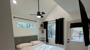 A bedroom with a bed, a ceiling fan, a window, a door, and a small shelf with a plant.