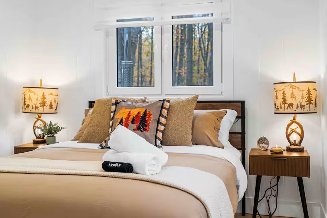 A neatly made bed with a blanket, pillows, and a folded towel. There are two bedside tables with lamps and a small plant.