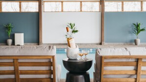 Zona de piscina con sillas de madera, una mesa de mármol con una botella y vasos, y plantas en macetas.