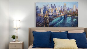 Ein Schlafzimmer mit Stadtbildgemälde, einer Nachttischlampe, einer Pflanze und einem Bett mit blauen und gelben Kissen.