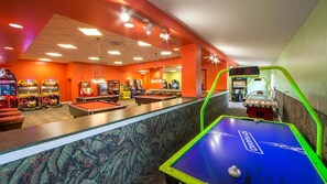 Una mesa de billar, juegos de arcade y una mesa de fútbolín en una sala luminosa con paredes anaranjadas.