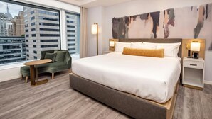 Une chambre d’hôtel moderne avec un grand lit, un fauteuil vert, une petite table et une vue sur la ville.