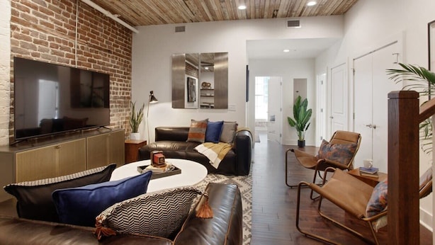 A living room with a brick wall, a flat-screen TV, leather sofas, a coffee table, and a wooden ceiling. - 10BR Downtown Savannah Combo | Walk to Nightlife (Savannah)