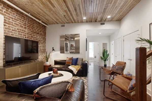 A living room with a brick wall, a flat-screen TV, leather sofas, a coffee table, and a wooden ceiling. - 10BR Downtown Savannah Combo | Walk to Nightlife (Savannah)