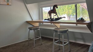 Ein Zimmer mit schrägverdeckter Decke, einem Fenster mit Blick auf Grünflächen, zwei Metallhocker und einem Holztisch.