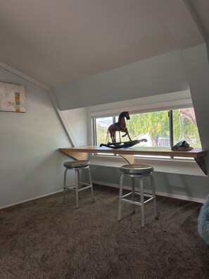 Ein Zimmer mit schrägverdeckter Decke, einem Fenster mit Blick auf Grünflächen, zwei Metallhocker und einem Holztisch.