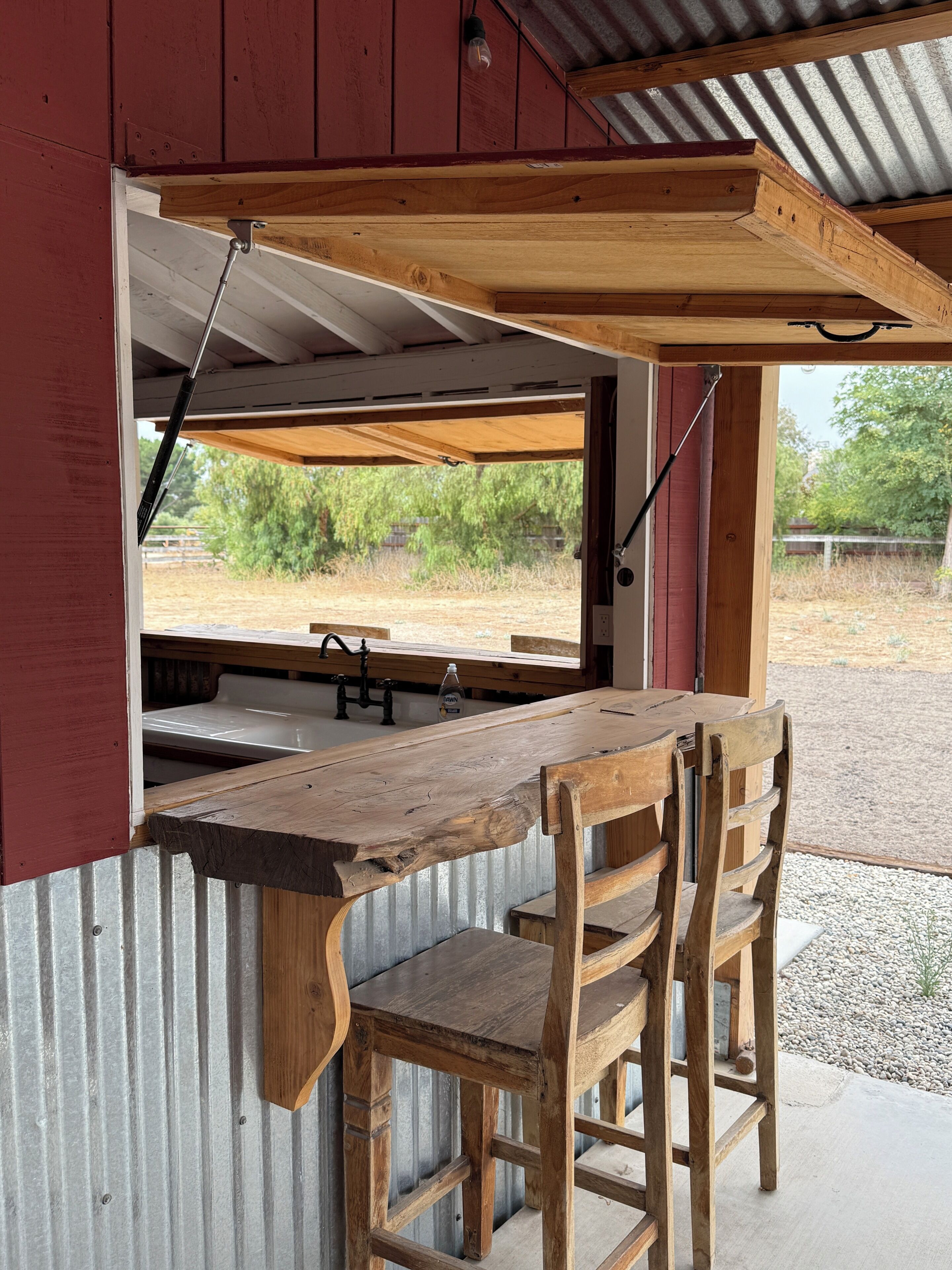 A rustic wooden bar with a metal sink and two wooden chairs, set against a corrugated metal wall with a view of a dry landscape.