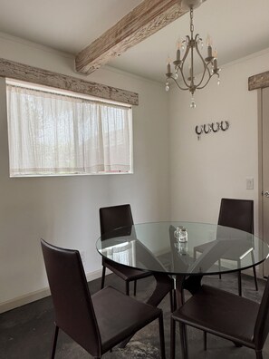 A dining area with a round glass table, four chairs, a chandelier, and a window with sheer curtains.