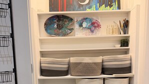 Uma lavanderia com máquina de lavar, prateleiras com cestos e um quadro na parede.