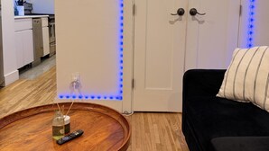 Quarto com piso de madeira, um sofá preto, uma mesa redonda de madeira com um controle remoto e uma porta branca com luzes LED azuis.