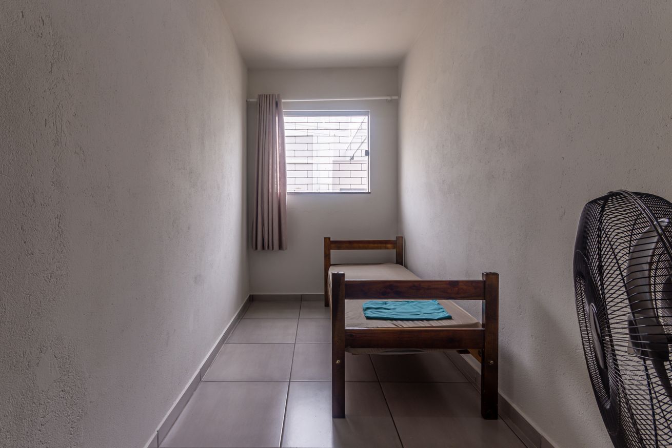 Une petite pièce avec un lit simple, une fenêtre avec un rideau et un ventilateur sur pied.