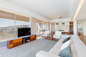 Una sala de estar moderna con un televisor grande, sofá y comedor, que cuenta con ventanas amplias y un balcón.