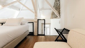 Ein Zimmer mit hohen Decken und sichtbarer Holzbalkenstruktur, einem Bett, einem Sessel und Blick durch ein Fenster.