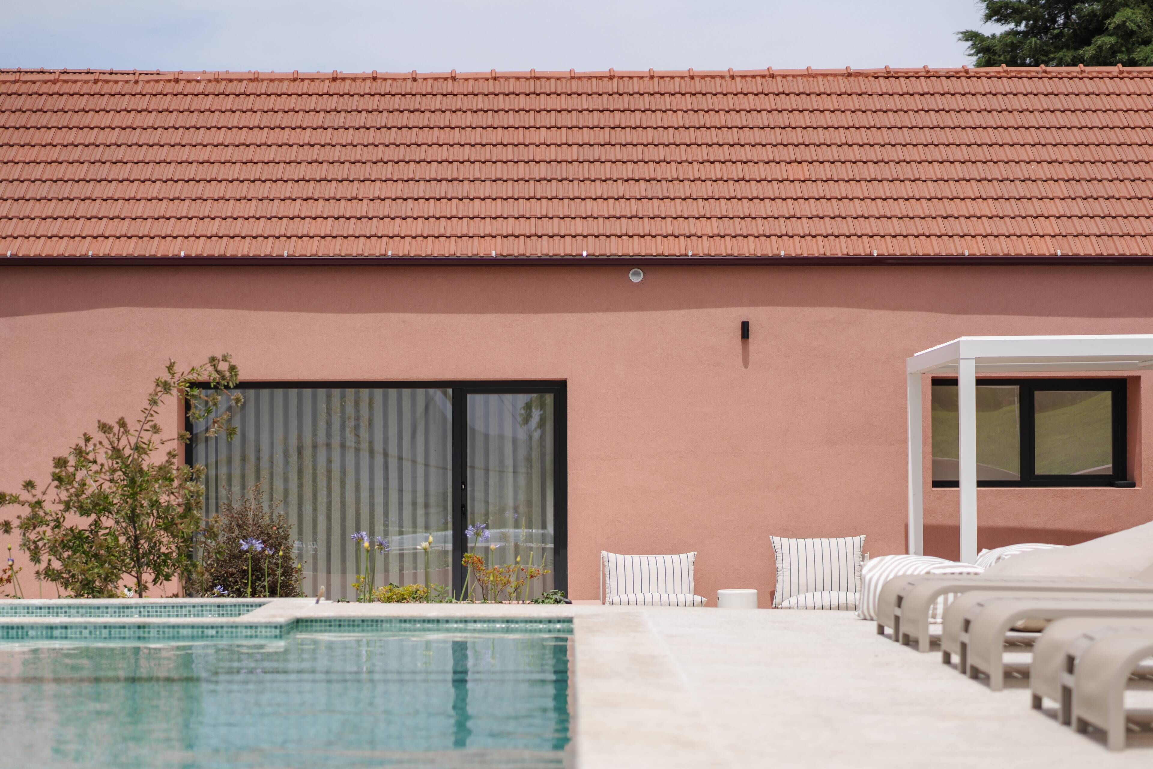 Un espace aménagé au bord de la piscine, avec des chaises longues, un coin salon abrité et un bâtiment doté d’un toit en tuiles.