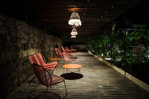 Un área de estar al aire libre con paredes de piedra, mesas y sillas, bajo un techo de madera con luces colgantes.