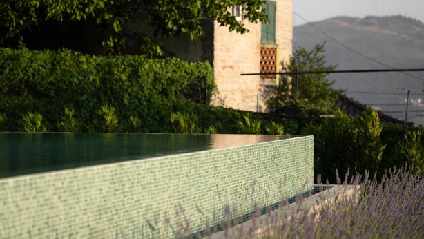 Ein Schwimmbecken mit einer gefliesten Kante, ein Steinhaus mit einem grünen Fenster und ein Berg im Hintergrund.