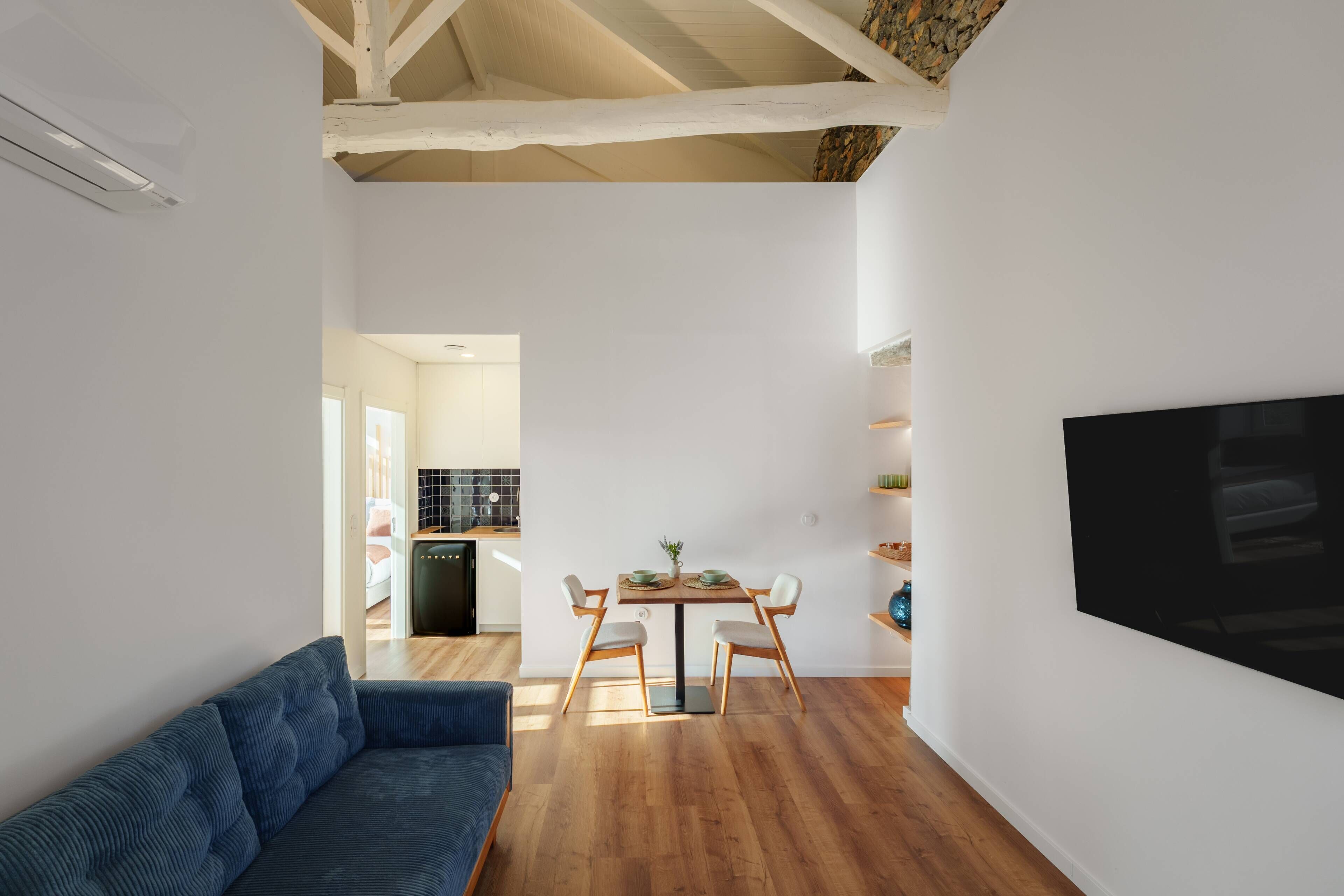 A modern living room with a sofa, dining table, and chairs, a kitchen in the background, and a TV mounted on the wall.