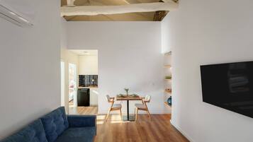 A modern living room with a sofa, dining table, and chairs, a kitchen in the background, and a TV mounted on the wall.