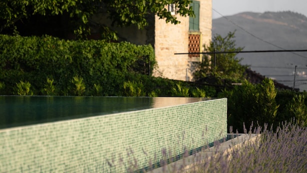 Une piscine bordée de carreaux, un bâtiment en pierre avec une fenêtre verte, et une montagne en arrière-plan.