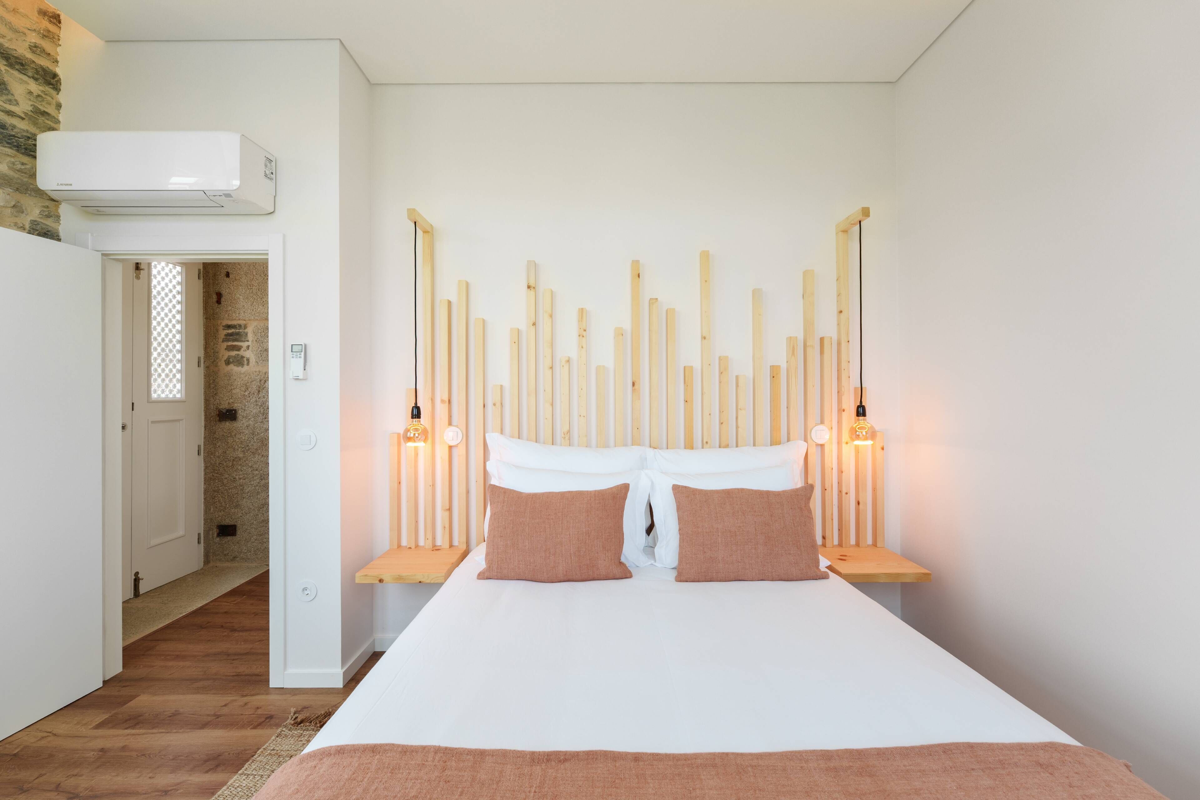 Quarto com cama, cabeceira de madeira, luminárias de cabeceira e ar condicionado fixado na parede.