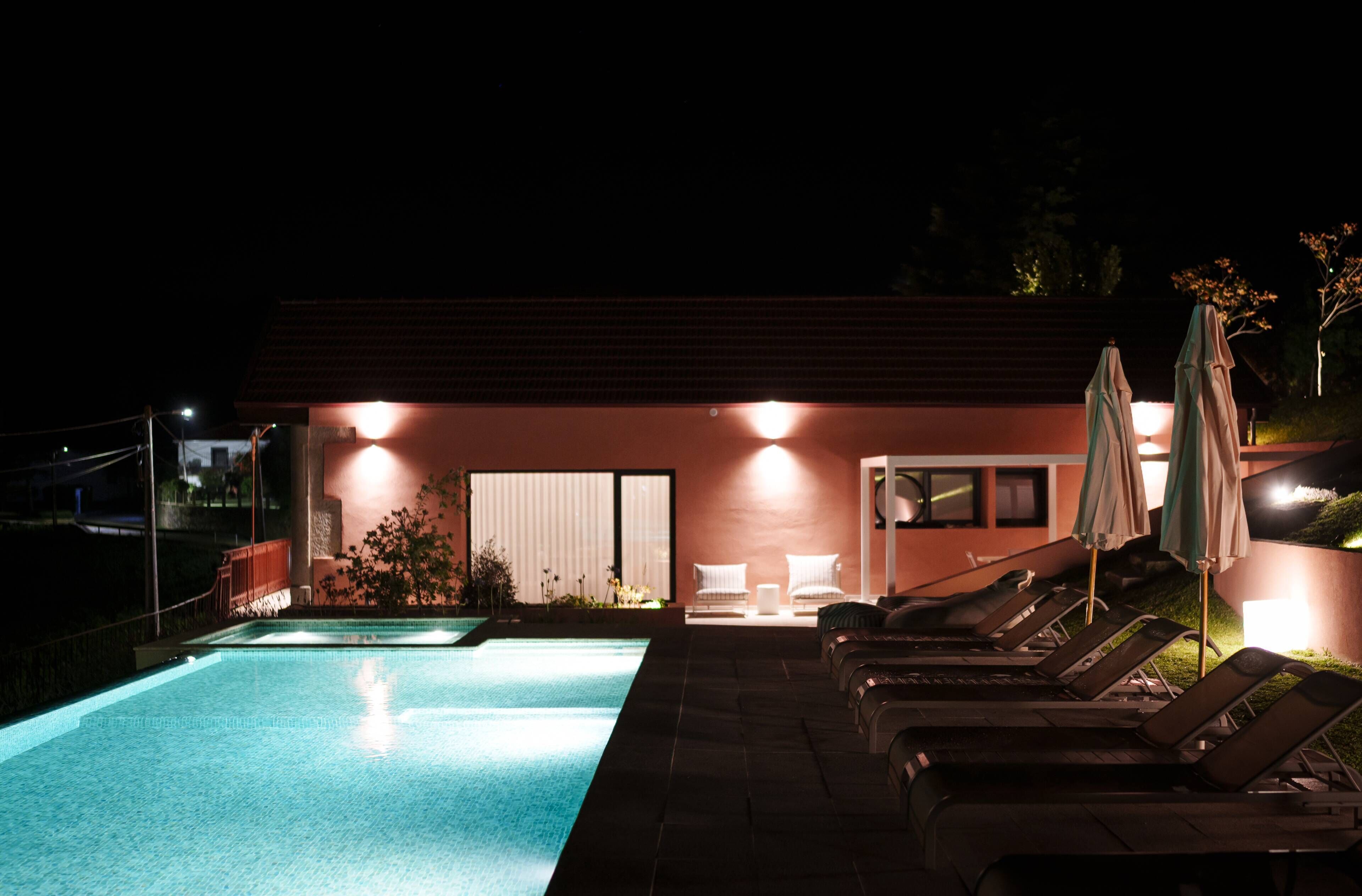 Une piscine avec des chaises longues et des parasols, un bâtiment à la façade rouge et une porte avec un volet blanc.