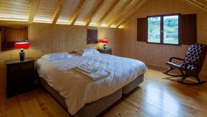 A bedroom with a wooden ceiling, a bed, two bedside tables with lamps, a rocking chair, and a window. - Casa do Alambique in Quinta da Bemposta (Vila Nova de Famalicão)