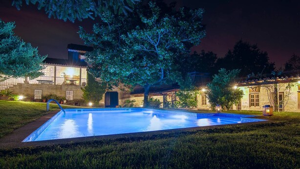 A swimming pool with a fountain, surrounded by well-lit buildings and trees. - Casa do Alambique in Quinta da Bemposta (Vila Nova de Famalicão)