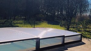 Piscina com cobertura de vidro, rodeada por um deck de madeira e árvores.