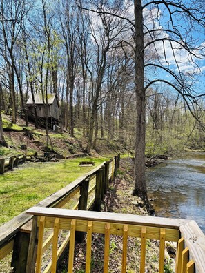 A wooden deck overlooking a river, surrounded by trees and a small cabin. - Serene 2-bedroom cottage along Marsh Creek in charming Gettysburg (Gettysburg)