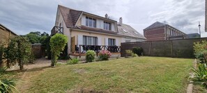 Ein zweistöckiges Haus mit geneigtem Dach, einem umzäunten Hinterhof und einem kleinen Garten mit Blumen.