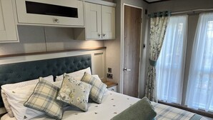 A neatly made bed with a headboard, pillows, and a rolled towel. There is a wooden door and a window with curtains. - Seafront at the Beach House at 248 Lydstep Haven Tenby Pembrokeshire (Tenby)