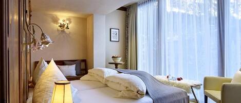 A hotel room with a bed, a bedside lamp, a chair, a window with curtains, and a wall-mounted light fixture.