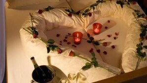 Un lit avec du linge de lit blanc, une table de chevet avec une bouteille et des verres, et une lampe fixée au mur.