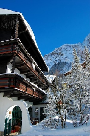 A traditional chalet with a sloped roof, wooden balconies, and a snow-covered mountain backdrop.