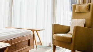 Une chambre avec un lit, une table de chevet en bois, un fauteuil vert et une vue sur les montagnes à travers de fines rideaux.
