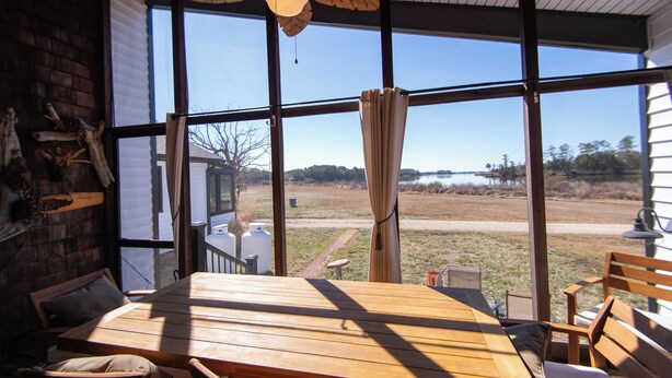 Une table et des chaises en bois dans une pièce dotée de grandes fenêtres, d’un ventilateur de plafond et offrant une vue sur un lac et des arbres.