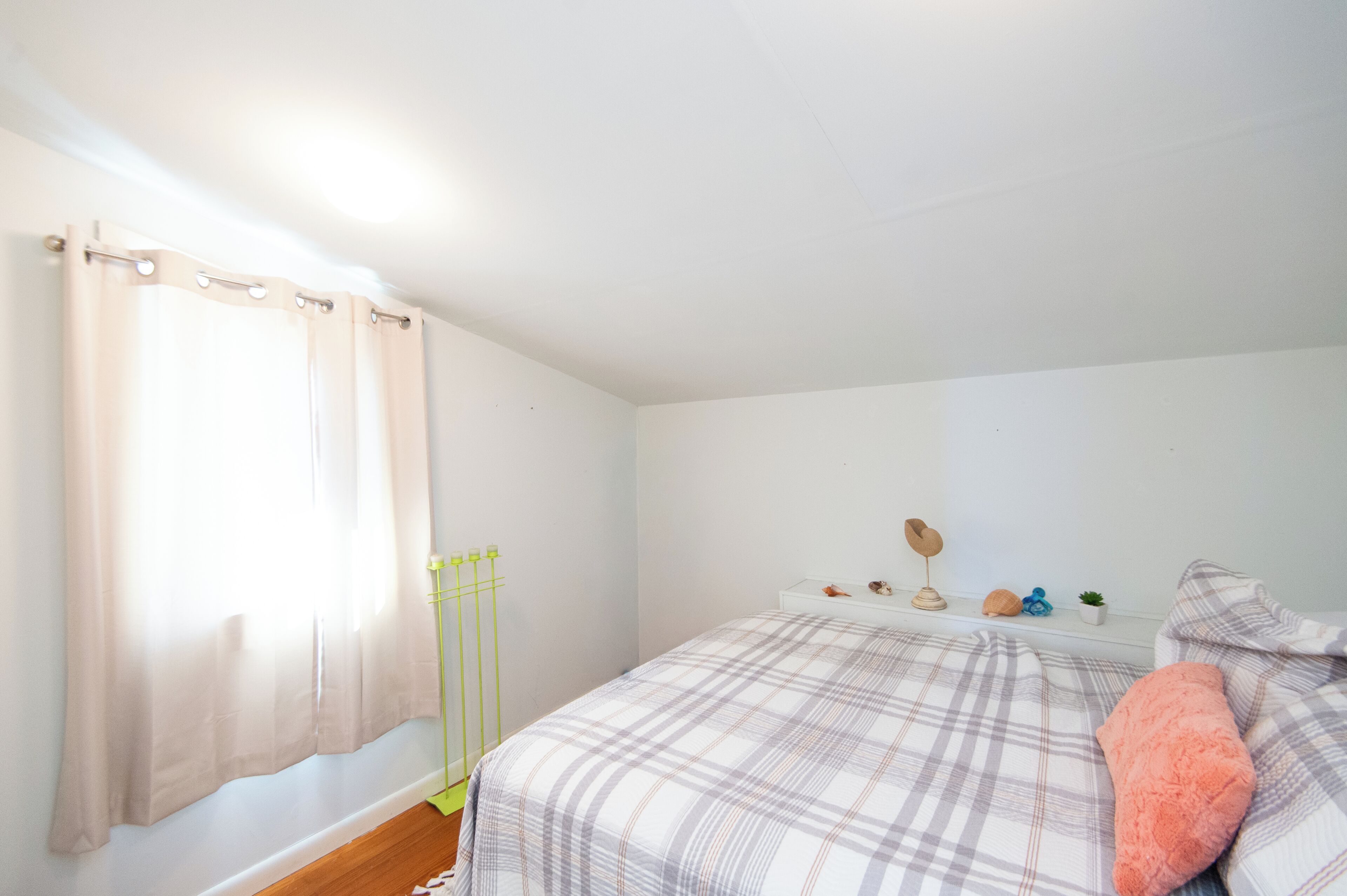Ein Schlafzimmer mit Bett, einem Fenster mit Vorhängen, einem Nachttisch mit Dekoration und einem an der Wand befestigten Regal.