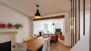 A dining area with a wooden table and chairs, a hanging light fixture, and a view into another room with a television.