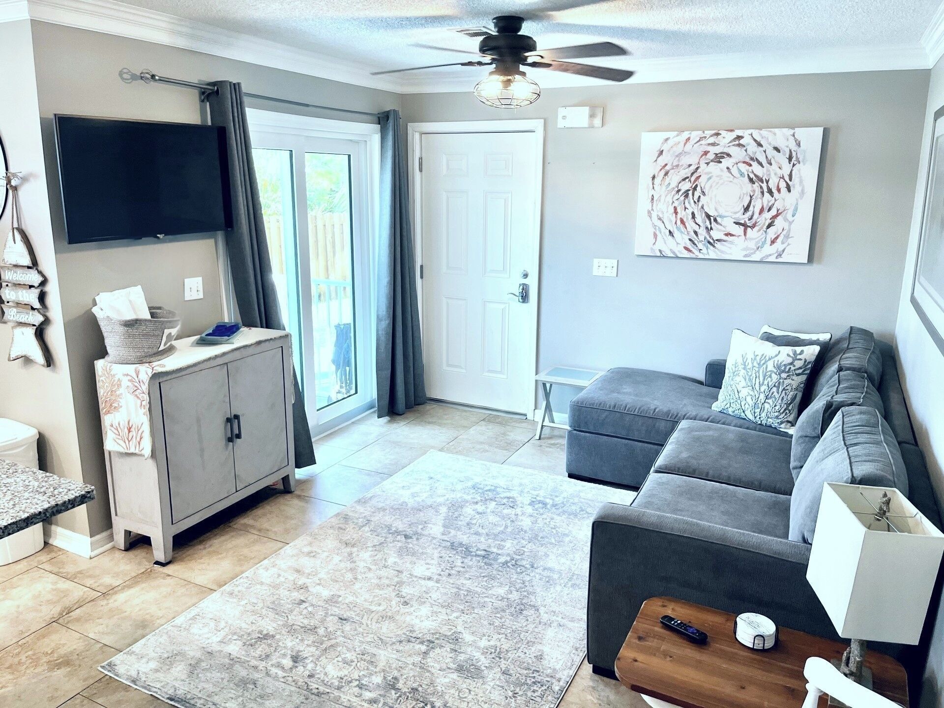 A living room with a gray sofa, a wooden coffee table, a TV mounted on the wall, and a door leading to a balcony.