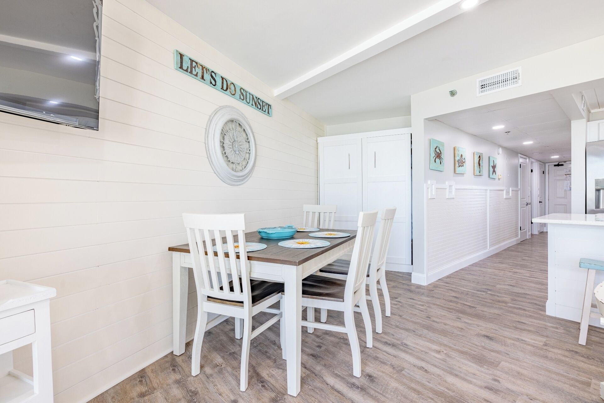 Un coin repas avec une table en bois et des chaises blanches, un élément décoratif mural et un panneau sur lequel on peut lire : « PROFITONS DU COUCHER DE SOLEIL ».