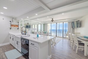 A modern kitchen with a central island, white cabinetry, and a dishwasher. A dining area with a table and chairs is visible, overlooking a balcony with a view of the ocean. - Sandy Sunsets Tower 3 - 6th floor, gorgeously updated beachfront w/2 king beds+ (Panama City Beach)