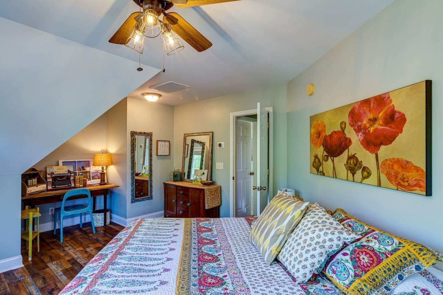 A bedroom with a sloped ceiling, a desk with a chair, a bed with patterned bedding, and a large floral painting on the wall.