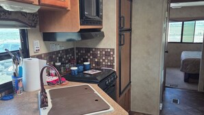 A compact RV kitchen with a microwave, stove, sink, and a window providing a view of the outdoors.