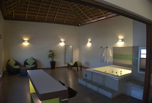 A spacious room with a large bathtub, seating area with chairs, a potted plant, and wall-mounted lights.