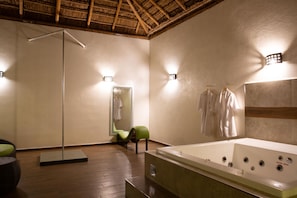 A spa room with a wooden floor, a large mirror, a green chair, a bathtub, and two wall-mounted lights.