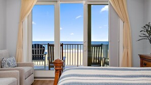 Une chambre à coucher comprenant un lit, une chaise, un canapé et donnant sur la plage grâce à une porte-fenêtre coulissante.