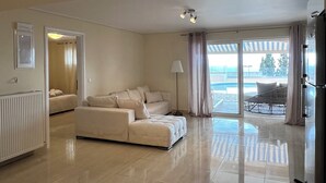 A spacious living room with a large sofa, a dining area with a table and chairs, and a view of a pool through the glass doors. - Seaview Villa Lagonissi |7-min Walk to Beach| Pool & SunsetViews |Athens Riviera (Kalivia Thorikou)