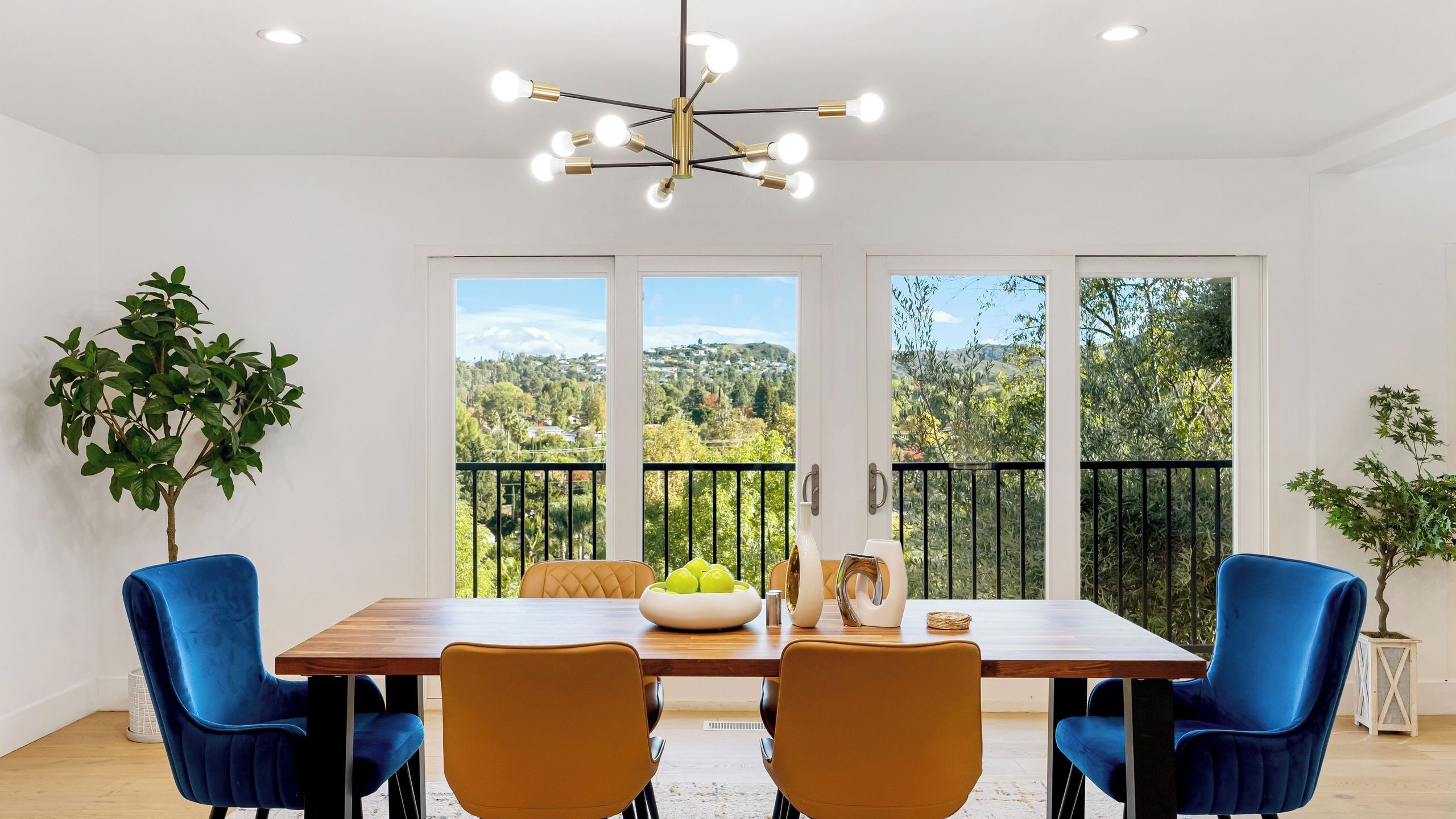 A dining area with a wooden table, blue and orange chairs, a chandelier, and a balcony with a view of greenery and hills.