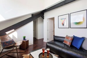 A loft-style living room with a black leather sofa, a rocking chair, and a coffee table with two glasses of beer. There are framed artworks on the wall and a staircase with exposed beams. - 6BR Downtown Savannah Condo | Walk to Nightlife (Savannah)